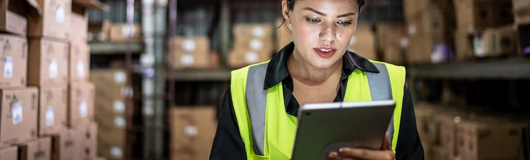 Frau mit gelber Warnweste, im Hintergrund ein Lager zu sehen, mit Tablet in der Hand. 